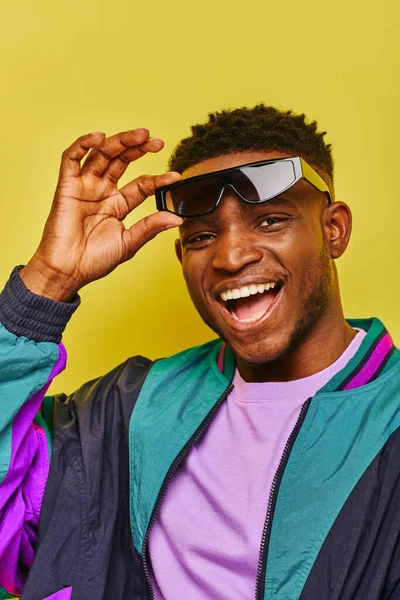 Unbekümmerter afrikanisch-amerikanischer Typ mit trendiger Sonnenbrille und farbenfroher Jacke blickt in die Kamera auf gelb — Stockfoto