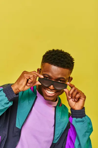 Cool homme afro-américain dans des lunettes de soleil à la mode et veste colorée souriant à la caméra sur jaune — Photo de stock