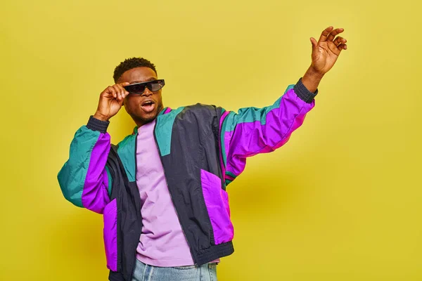 Excité homme afro-américain en lunettes de soleil et veste colorée à la mode agitant la main sur jaune — Photo de stock