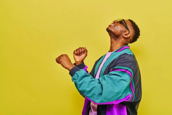 Young and trendy african american man in colorful jacket and sunglasses dancing on yellow — Stock Photo