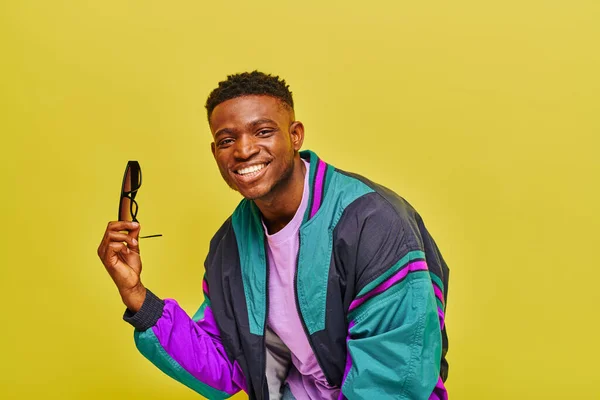 Handsome african american man in colorful jacket holding sunglasses and smiling at camera on yellow — Stock Photo