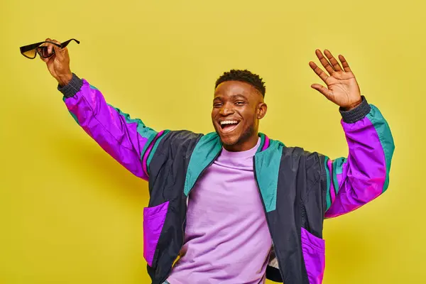 Excited african american man in bright jacket holding sunglasses and dancing on yellow backdrop — Stock Photo