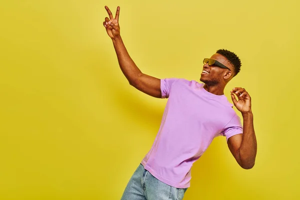 Uomo afroamericano sorridente in eleganti occhiali da sole e t-shirt viola che mostra il segno della vittoria sul giallo — Foto stock