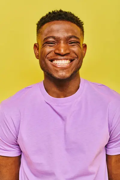 Homme afro-américain joyeux en t-shirt violet avec sourire radieux en regardant la caméra sur fond jaune — Photo de stock