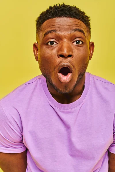 Amazed african american man in purple t-shirt with open mouth looking at camera on yellow backdrop — Stock Photo