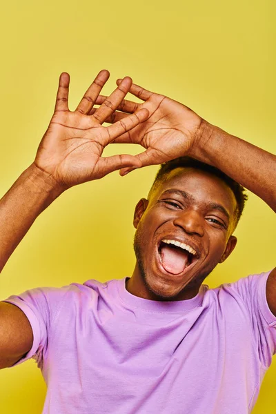 Alegre y divertido afroamericano hombre gestos y sobresaliendo lengua en amarillo telón de fondo - foto de stock