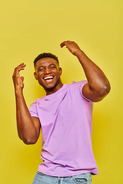 Схвильований і модний афроамериканський чоловік у фіолетовій футболці жестикулює сміючись на жовтому тлі — стокове фото