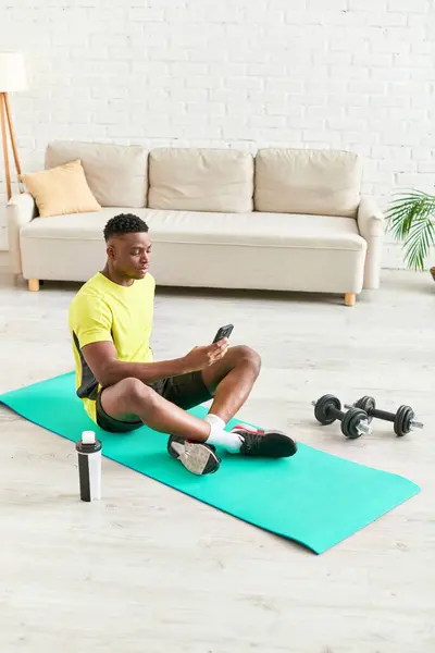 Joven afroamericano hombre en ropa deportiva sentado en la alfombra de fitness con teléfono inteligente cerca de pesas - foto de stock