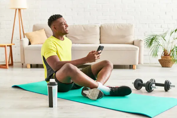 Chico afroamericano feliz en ropa deportiva sentado en la alfombra de fitness con teléfono inteligente cerca de pesas - foto de stock
