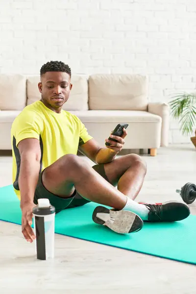 Chico afroamericano feliz en ropa deportiva sentado en la alfombra de fitness con teléfono inteligente cerca de pesas - foto de stock