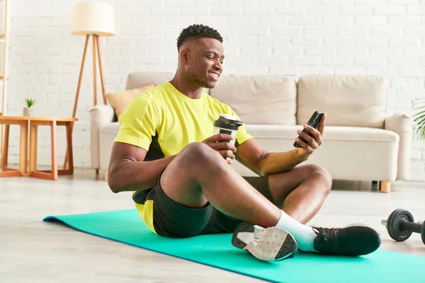 African american man in sportswear holding smartphone taking sports bottle on fitness mat at home — Stock Photo