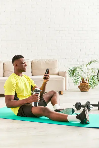Hombre americano africano alegre en ropa deportiva sentado en la estera de la aptitud con la botella de deportes y el teléfono inteligente - foto de stock
