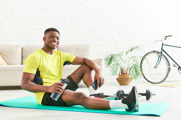 Hombre afroamericano sonriente en ropa deportiva con estilo en la alfombra de fitness con botella de deportes y teléfono inteligente - foto de stock