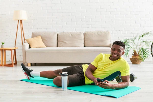Alegre afroamericano hombre con teléfono inteligente mirando a la cámara en la estera de fitness en la sala de estar - foto de stock