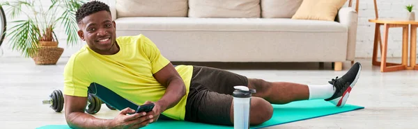 Sonriente hombre negro con teléfono inteligente mirando a la cámara en la alfombra de fitness cerca de la botella de deportes, pancarta - foto de stock