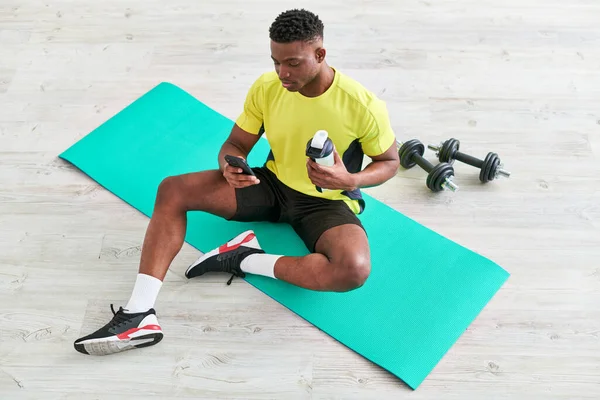 Fit uomo afroamericano in abbigliamento sportivo seduto su tappetino fitness con smartphone e bottiglia sportiva — Foto stock