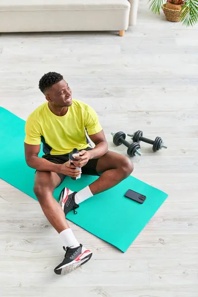 Щасливий афроамериканський чоловік у спортивному одязі, що сидить на фітнес-матусі зі спортивною пляшкою біля смартфона — стокове фото