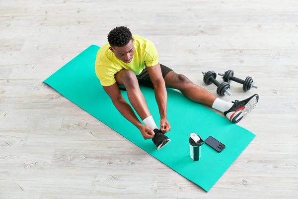 Молодой африканский американец шнурует кроссовки на коврике для фитнеса рядом со смартфоном и спортивным оборудованием — стоковое фото