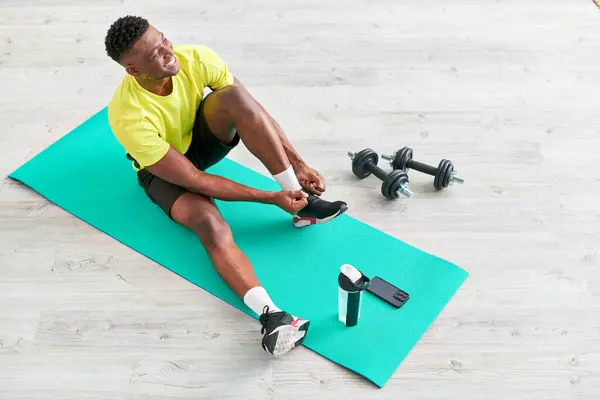Sonriente afroamericano hombre atando zapatilla de deporte en la alfombra de fitness cerca de teléfono inteligente y equipo deportivo - foto de stock
