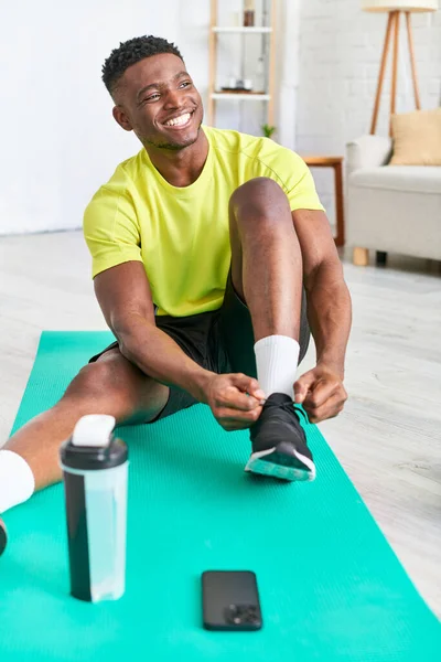 Chico afroamericano positivo atando zapatilla de deporte en la estera de fitness cerca de teléfono inteligente y botella de deportes - foto de stock