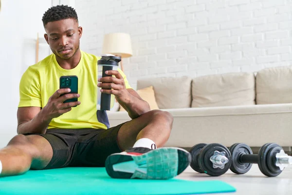 Joven afroamericano hombre con botella deportiva navegar por Internet en el teléfono inteligente en la alfombra de fitness en casa - foto de stock