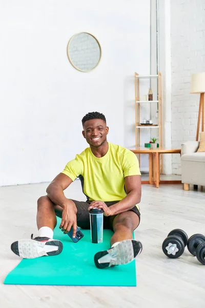 Despreocupado hombre afroamericano con botella deportiva y teléfono móvil mirando a la cámara en la colchoneta de fitness - foto de stock