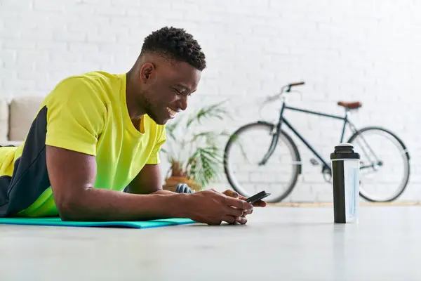 Alegre deportista afroamericano hombre con teléfono inteligente acostado en la colchoneta de fitness cerca de la botella de deportes - foto de stock