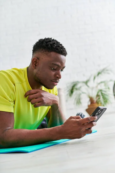 Hombre afroamericano serio que navega por las redes sociales en el teléfono inteligente mientras está acostado en la colchoneta de fitness - foto de stock