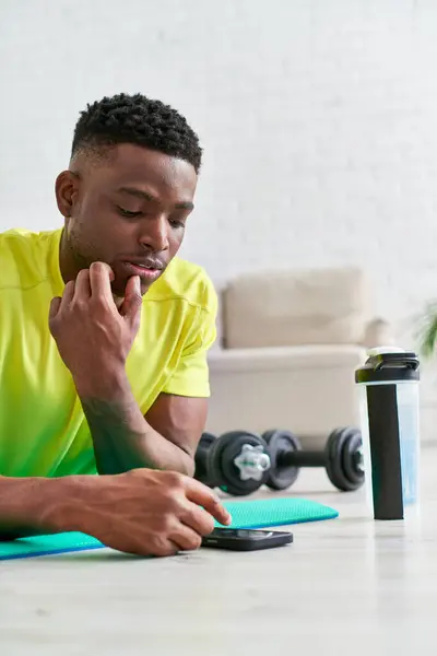 Reflexivo afroamericano hombre navegar por Internet en el teléfono inteligente mientras se acuesta en la colchoneta de fitness - foto de stock