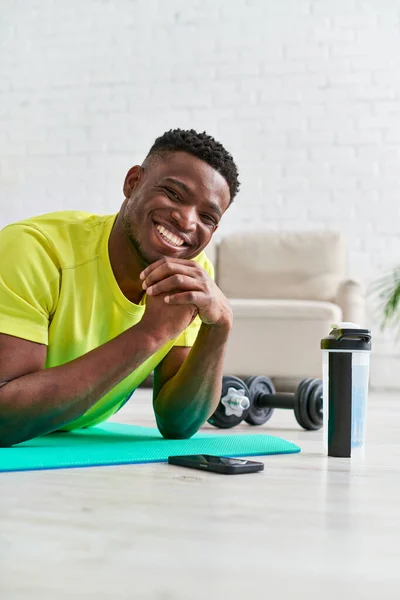 Feliz afroamericano sonriendo a la cámara en la colchoneta de fitness cerca de teléfono inteligente y botella de deportes en casa - foto de stock