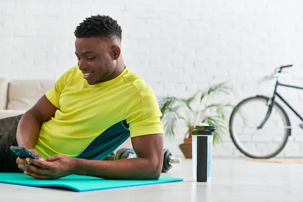 Sonriente hombre afroamericano con teléfono inteligente navegar por Internet en la alfombra de fitness cerca de la botella de deportes - foto de stock