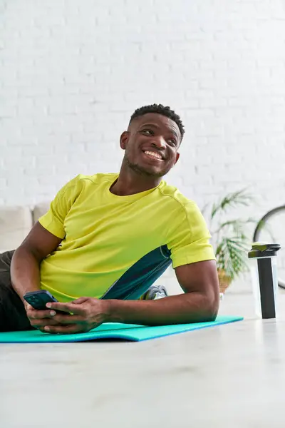 Alegre deportista afroamericano hombre con teléfono inteligente mirando hacia otro lado en la estera de fitness cerca de la botella de deportes - foto de stock