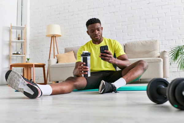 Ajuste hombre afroamericano con botella deportiva navegar por Internet en el teléfono inteligente sentado en la colchoneta de fitness - foto de stock