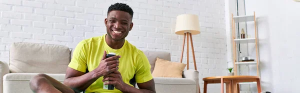 Glücklich sportlich afrikanisch-amerikanischer Mann mit Sportflasche blickt in die Kamera im modernen Wohnzimmer, Banner — Stockfoto
