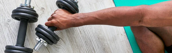 Vista parcial del hombre afroamericano deportivo sentado en la colchoneta de fitness y tomando mancuerna, pancarta - foto de stock