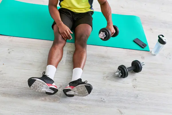 Vista cortada do atlético homem americano africano em sportswear sentado no tapete de fitness com haltere — Fotografia de Stock