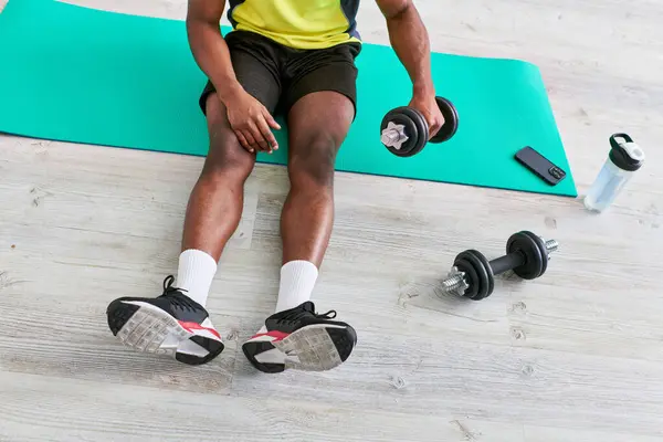 Recortado vista de atlético afroamericano hombre en ropa deportiva sentado en la alfombra de fitness con mancuerna - foto de stock