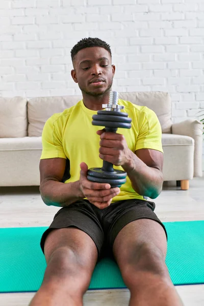 Joven atlético afroamericano hombre en ropa deportiva sentado en la alfombra de fitness y la celebración de la mancuerna - foto de stock