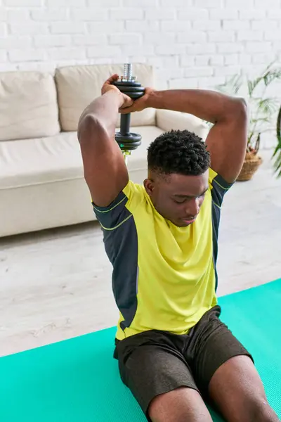 Atlético afroamericano hombre en ropa deportiva sentado en la alfombra de fitness y hacer ejercicio con la mancuerna - foto de stock
