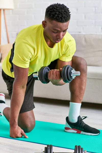 Atletico uomo afroamericano in abbigliamento sportivo che si allena con i manubri sul tappeto fitness a casa — Foto stock