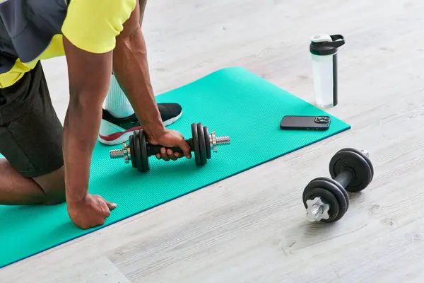 Hombre afroamericano recortado en ropa deportiva haciendo ejercicio con mancuerna en alfombra de fitness en casa - foto de stock