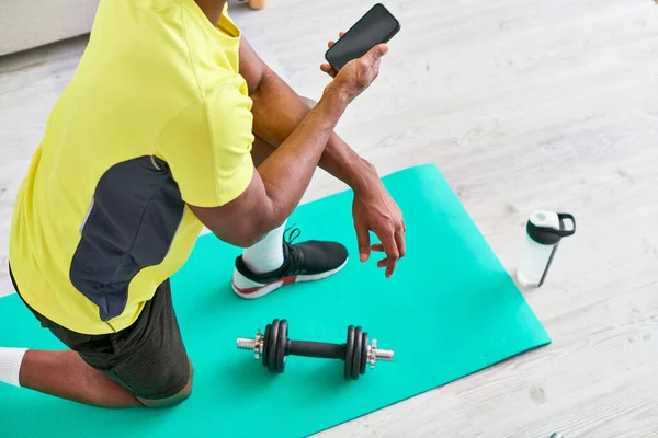 Hombre afroamericano recortado en ropa deportiva que sostiene el teléfono inteligente cerca de la mancuerna de la estera de fitness - foto de stock