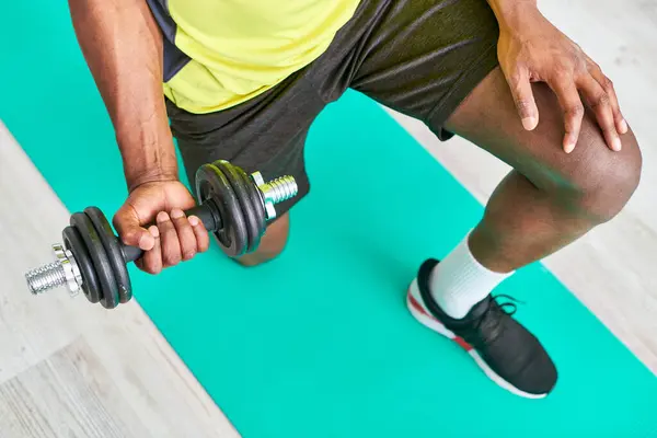 Vista recortada del hombre afroamericano en ropa deportiva haciendo ejercicio con mancuerna en la alfombra de fitness en casa - foto de stock