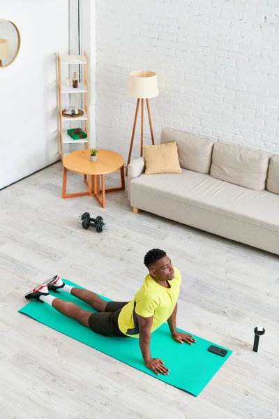Vista de ángulo alto del hombre afroamericano en ropa deportiva practicando la pose de yoga de serpiente en la estera de fitness - foto de stock