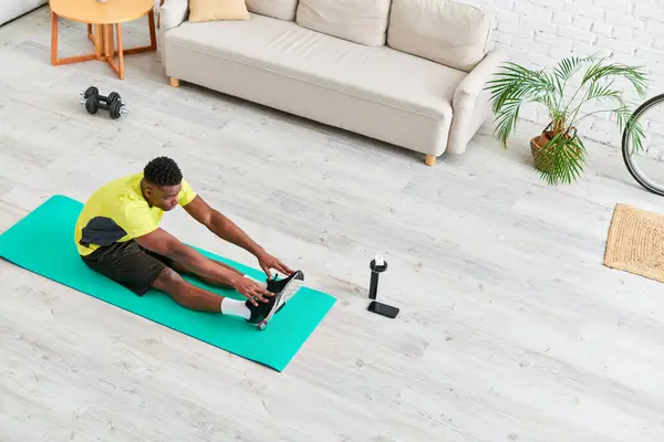 Hombre afroamericano en ropa deportiva que se extiende en la estera de fitness en la sala de estar moderna, vista de ángulo alto - foto de stock