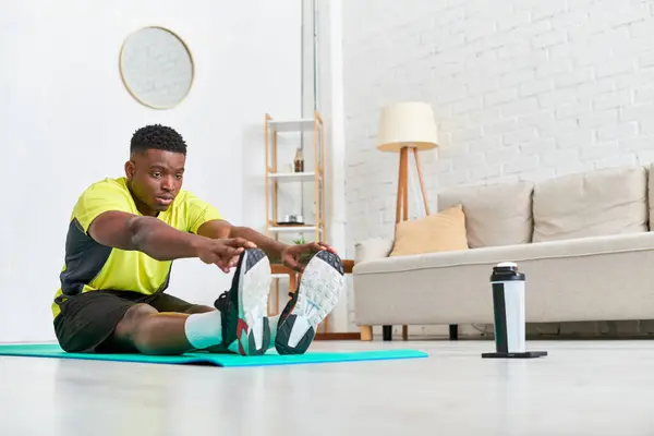Konzentrierter afrikanisch-amerikanischer Mann in Sportbekleidung, der sich im modernen Wohnzimmer auf einer Fitnessmatte dehnt — Stockfoto