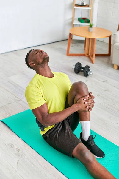 Atlético afroamericano chico sentado y estiramiento en el hombre de fitness en la sala de estar, entrenamiento en casa - foto de stock