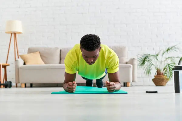 Hombre afroamericano en entrenamiento de ropa deportiva en postura de tablón en alfombra de fitness en la sala de estar moderna - foto de stock