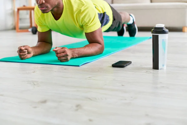 Hombre afroamericano cultivado entrenamiento en postura de tablón en la colchoneta de fitness cerca de la botella de deportes y teléfono inteligente - foto de stock