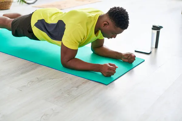 Sportif afro-américain guy formation en planche pose sur tapis de fitness près de bouteille de sport et smartphone — Photo de stock